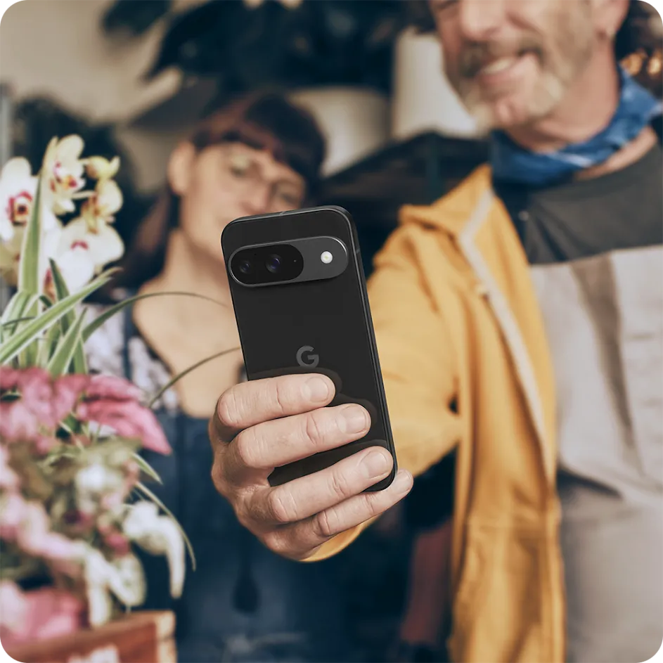 Photograph of a smiling middle-aged man taking a selfie with his Pixel phone, with a flower arrangement in the left foreground and a woman looking on over his shoulder in the background.